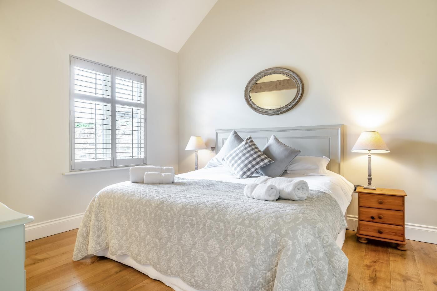 Quarto com uma cama grande, duas mesas de cabeceira, um espelho redondo e uma janela com persianas.
