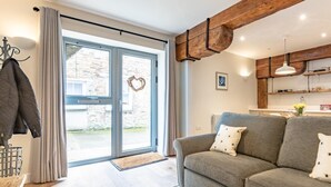 En moderne stue med en grå sofa, træbjælker, et køkken i baggrunden og en stor skydedør i glas.