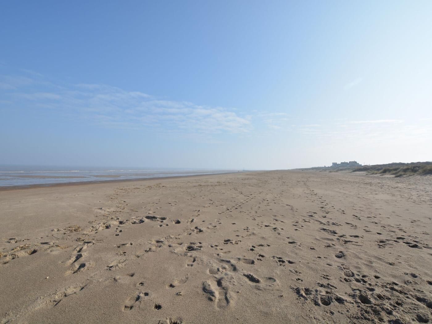 En bred sandstrand med fodaftryk, en klar himmel og fjerne klitter.