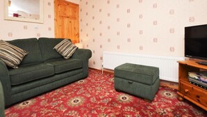 En stue med en grøn sofa, tilhørende skammel, et fjernsyn, en radiator og tapet med mønster.