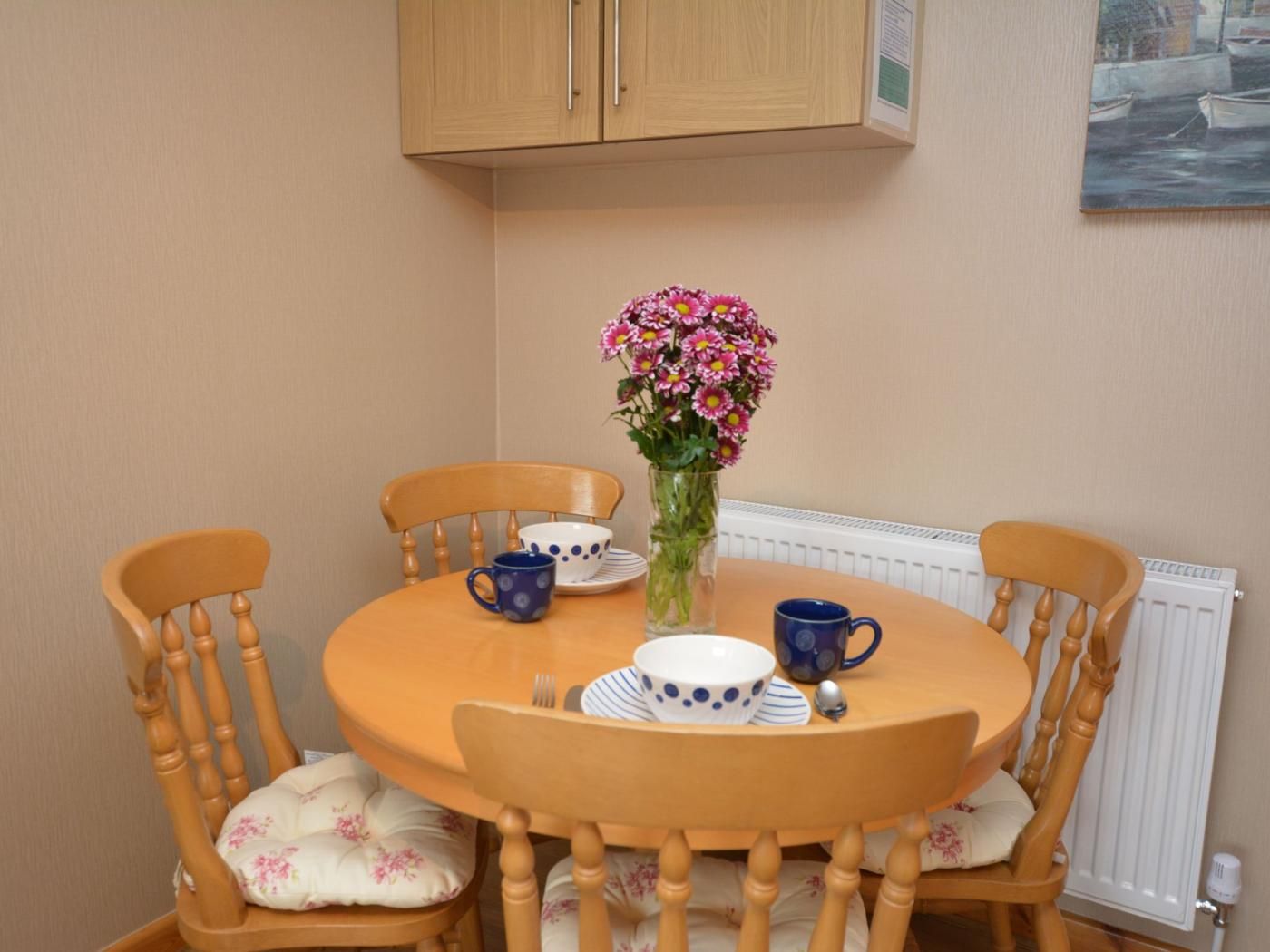 Spiseområde med et rundt bord, fire træstole, en vase med blomster og et billede på væggen.