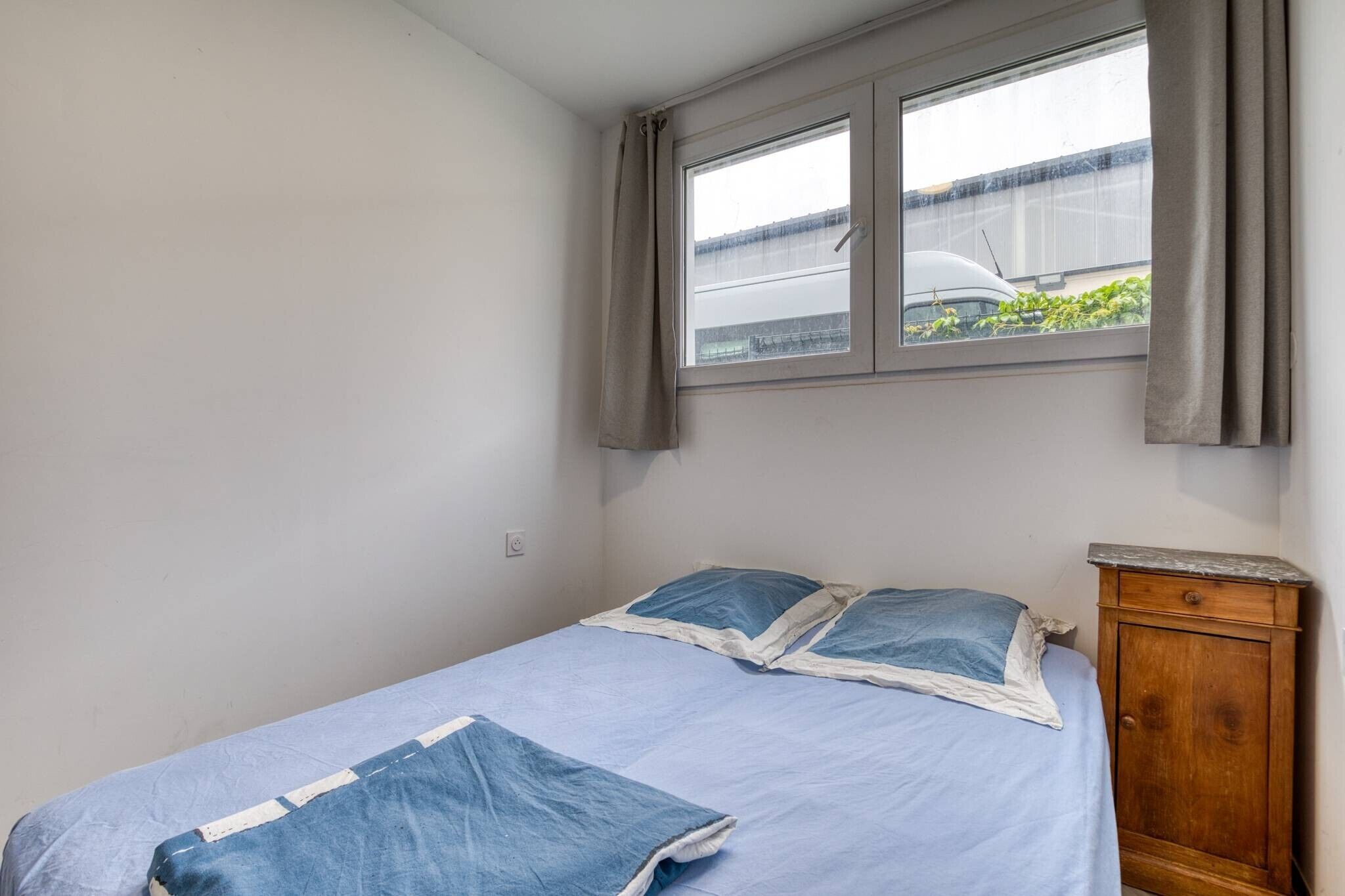Een enkel bed met een blauwe spreidek en kussens, een houten nachtkastje en een raam met gordijnen.