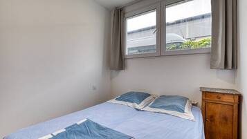 Een enkel bed met een blauwe spreidek en kussens, een houten nachtkastje en een raam met gordijnen.