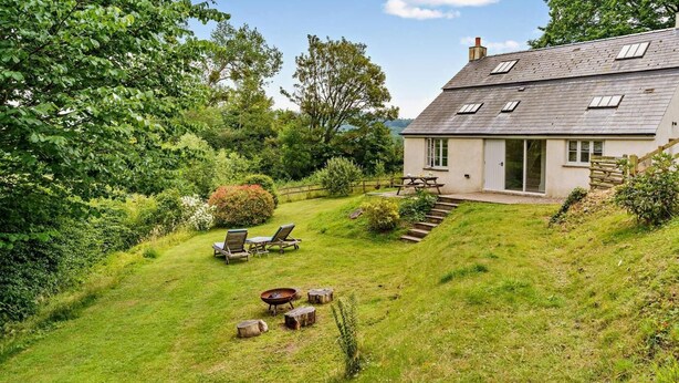 Ein Haus mit schrägem Dach, einer Terrasse mit Tisch und Stühlen sowie ein Garten mit einem kleinen Teich.