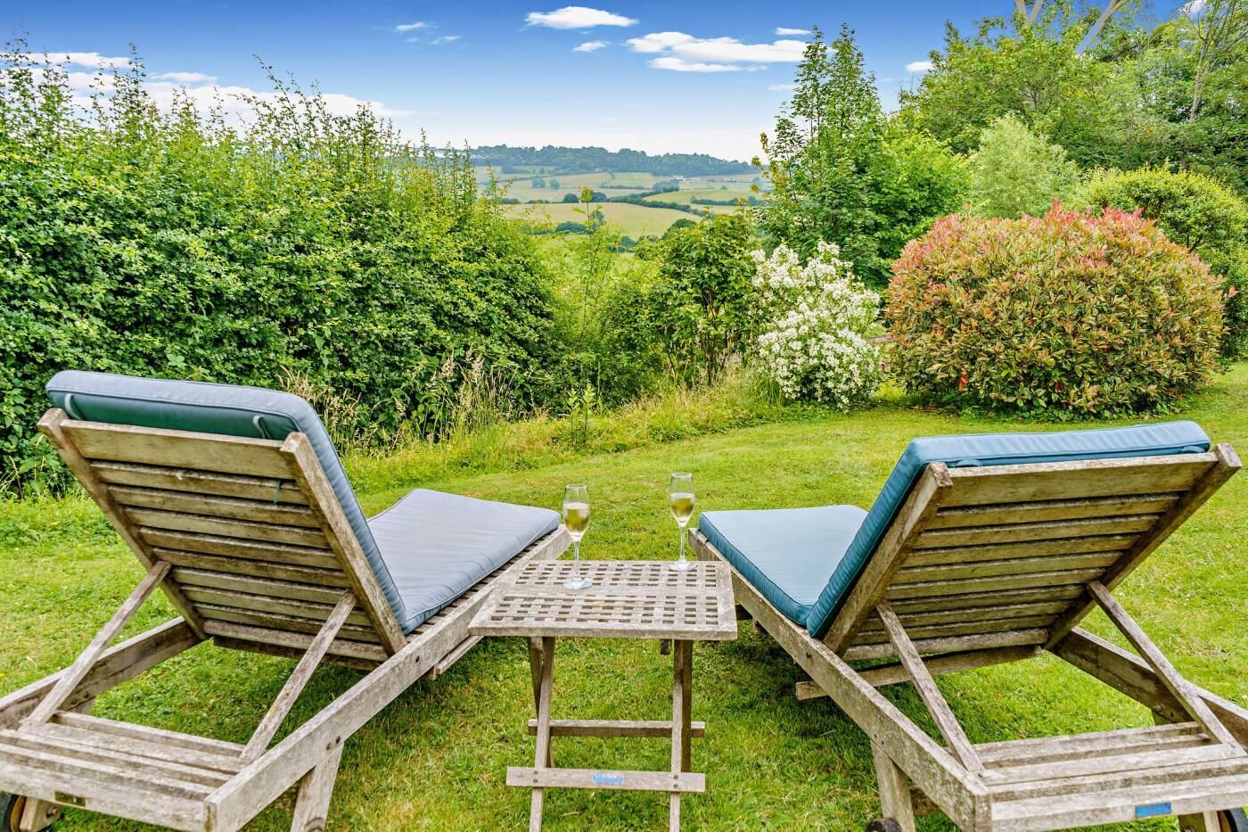 Two wooden lounge chairs with cushions, a small table, and a glass of wine, set against a backdrop of lush greenery and a distant view of rolling hills.