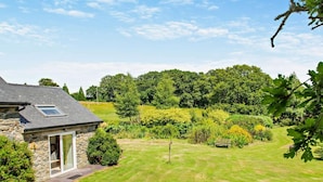 A stone house with a small attached structure, a well-maintained lawn, and dense forest in the background. - 2 Bed in Conwy (oc-42573) (Ty'n-y-groes)