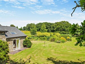 A stone house with a small attached structure, a well-maintained lawn, and dense forest in the background. - 2 Bed in Conwy (oc-42573) (Ty'n-y-groes)