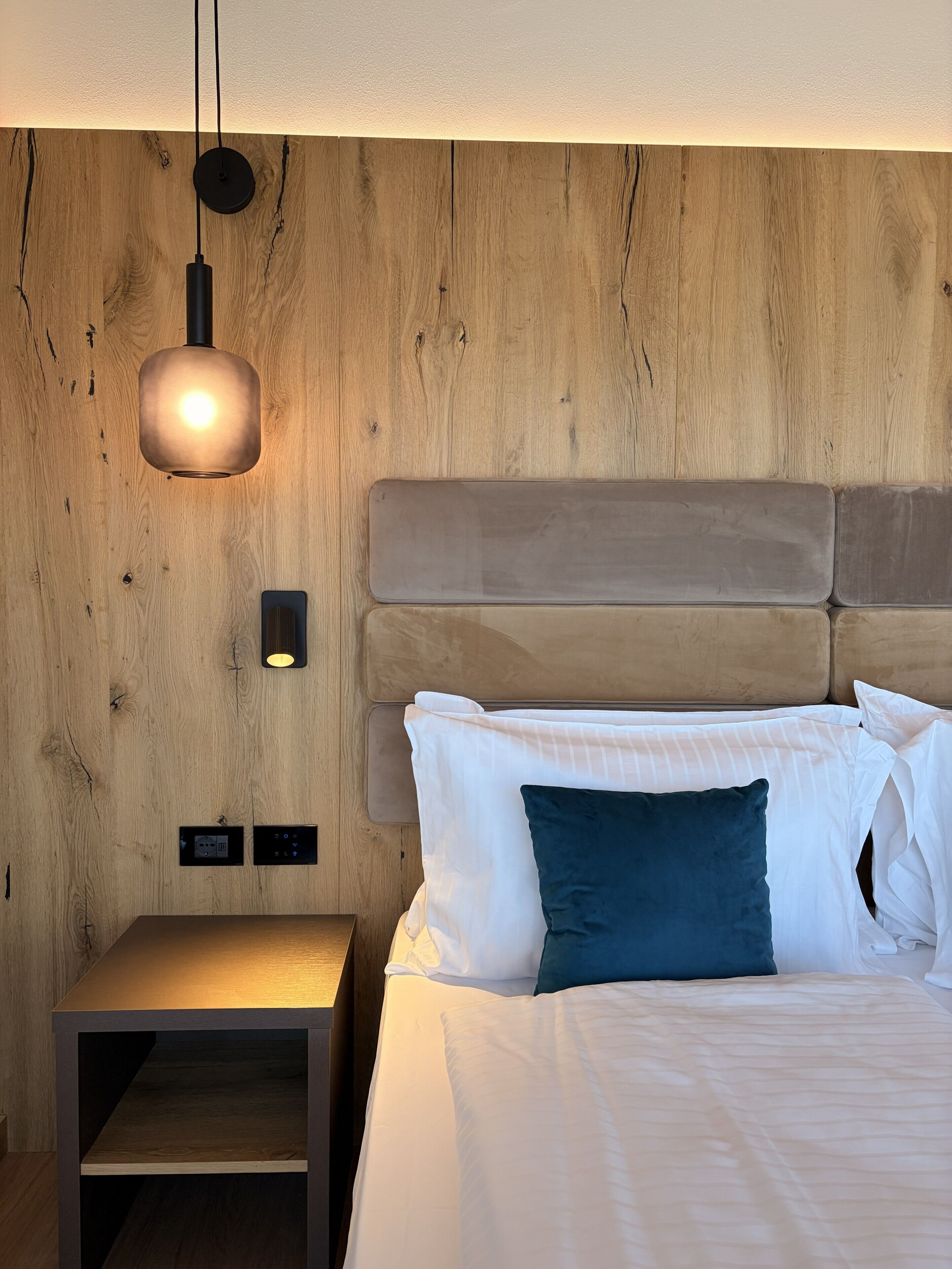 Une chambre avec un mur en bois, un lit avec des oreillers blancs et bleu foncé, une table de chevet et un luminaire suspendu.