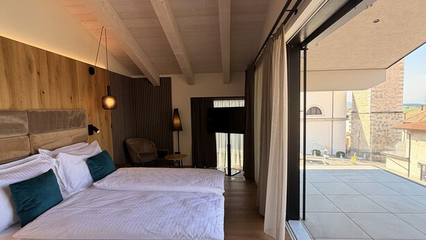 A modern hotel room with a large bed, a wooden headboard, a desk, and a view through a sliding glass door. - ALPEN RELAIS (Fondo)
