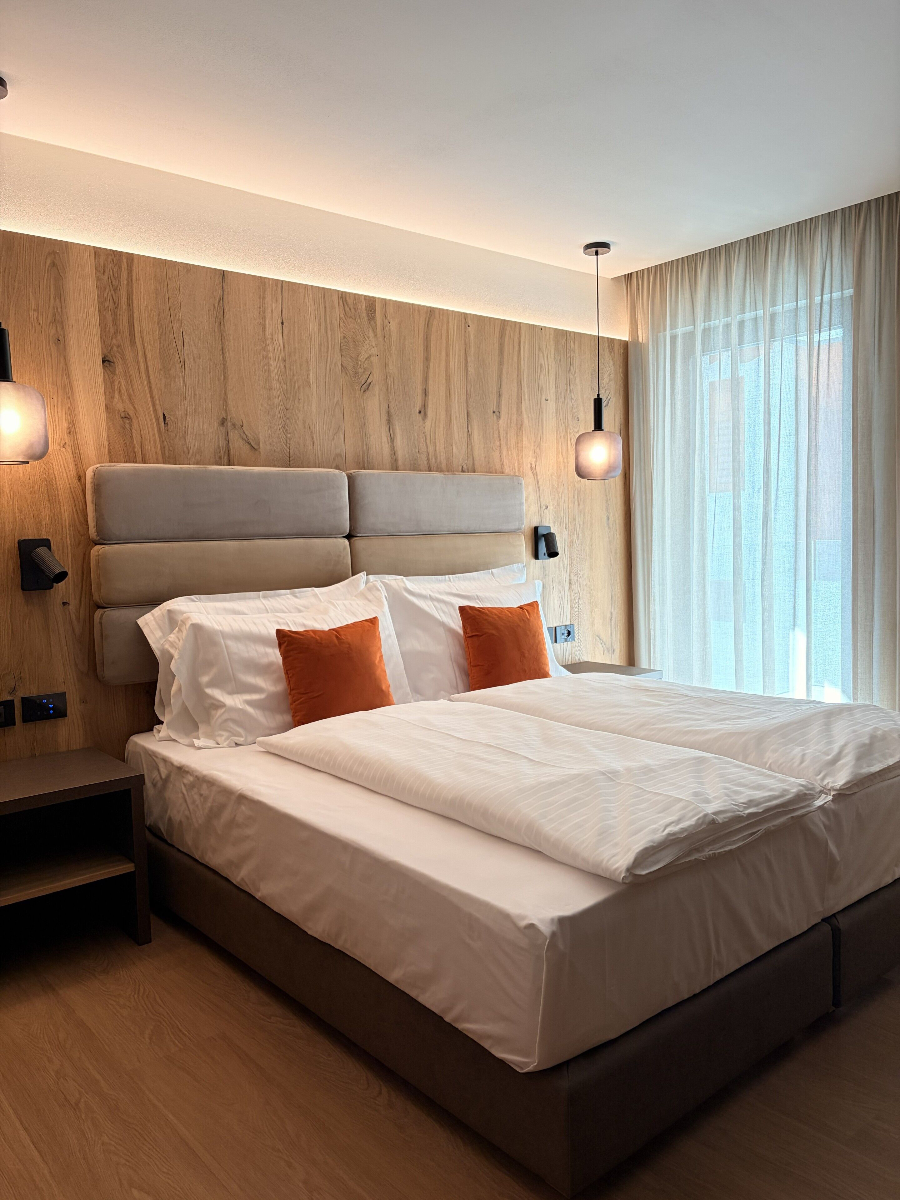 A modern hotel room with a wooden headboard, a large bed with white linens and orange pillows, bedside lamps, and a view through sheer curtains.