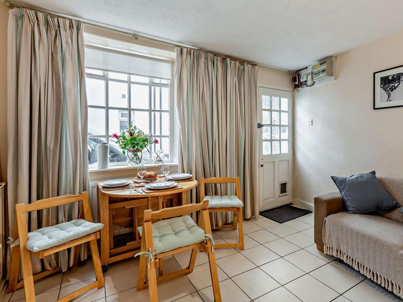 Um quarto com uma mesa de jantar posta para quatro pessoas, duas cadeiras de madeira, um sofá, uma janela com cortinas e uma porta.