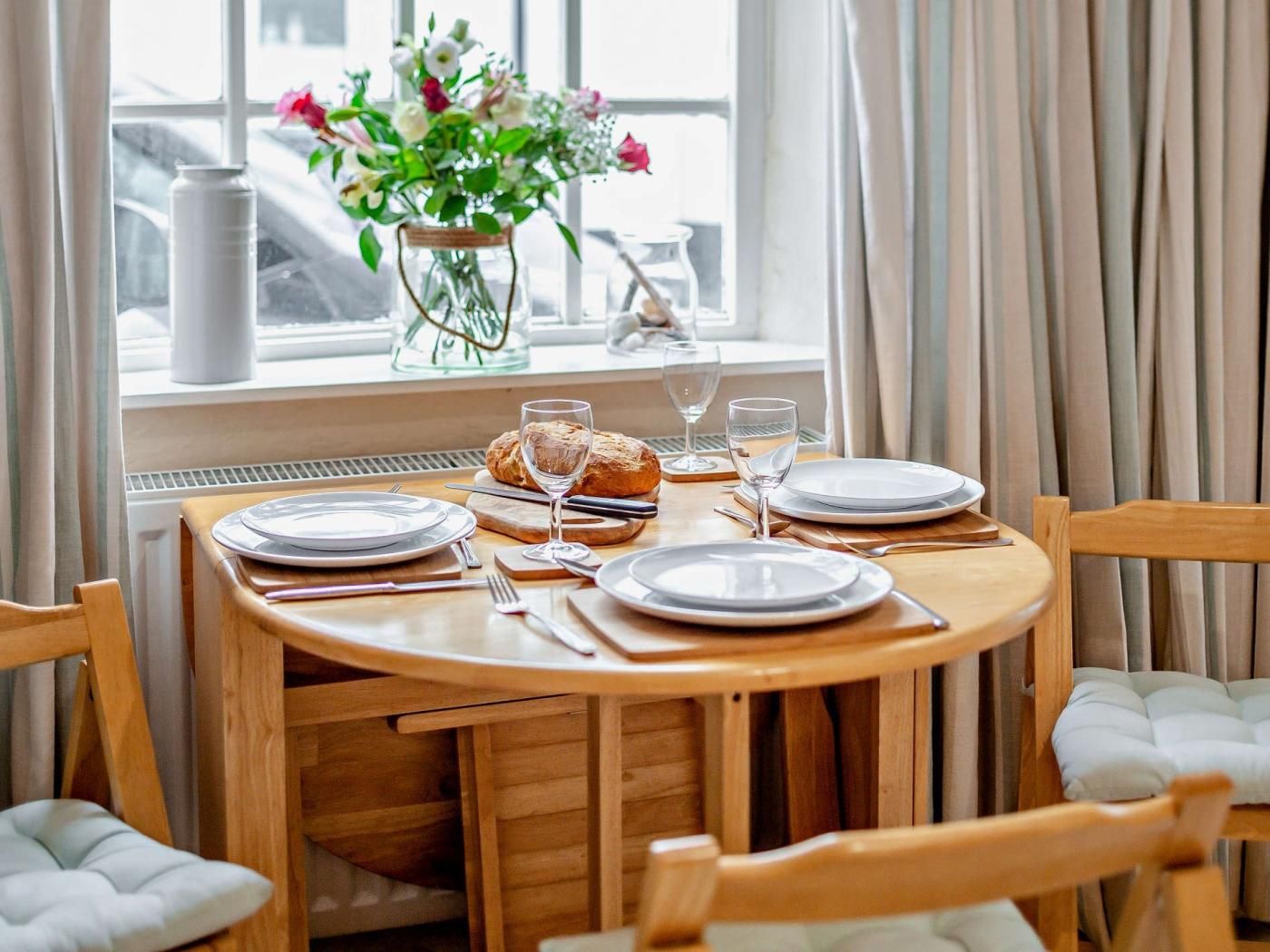 Una mesa de comedor dispuesta con platos, cubiertos, pan y un florero.