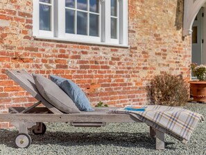 A wooden lounge chair with cushions, a book, and a plaid blanket on a gravel surface, against a brick wall with a window. - 8 Bed in Kington (oc-42962) (Titley)