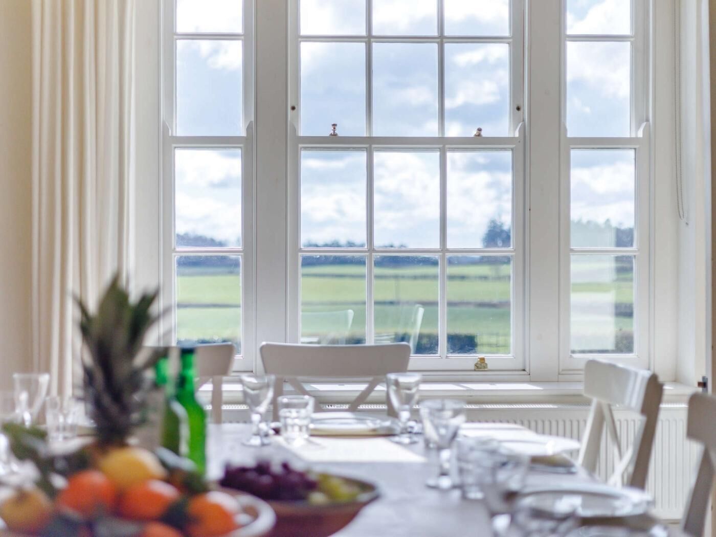 Ein Essbereich mit Tisch für zwei Personen, Blick auf ein grünes Feld durch große Fenster und eine Schale mit Obst auf dem Tisch.