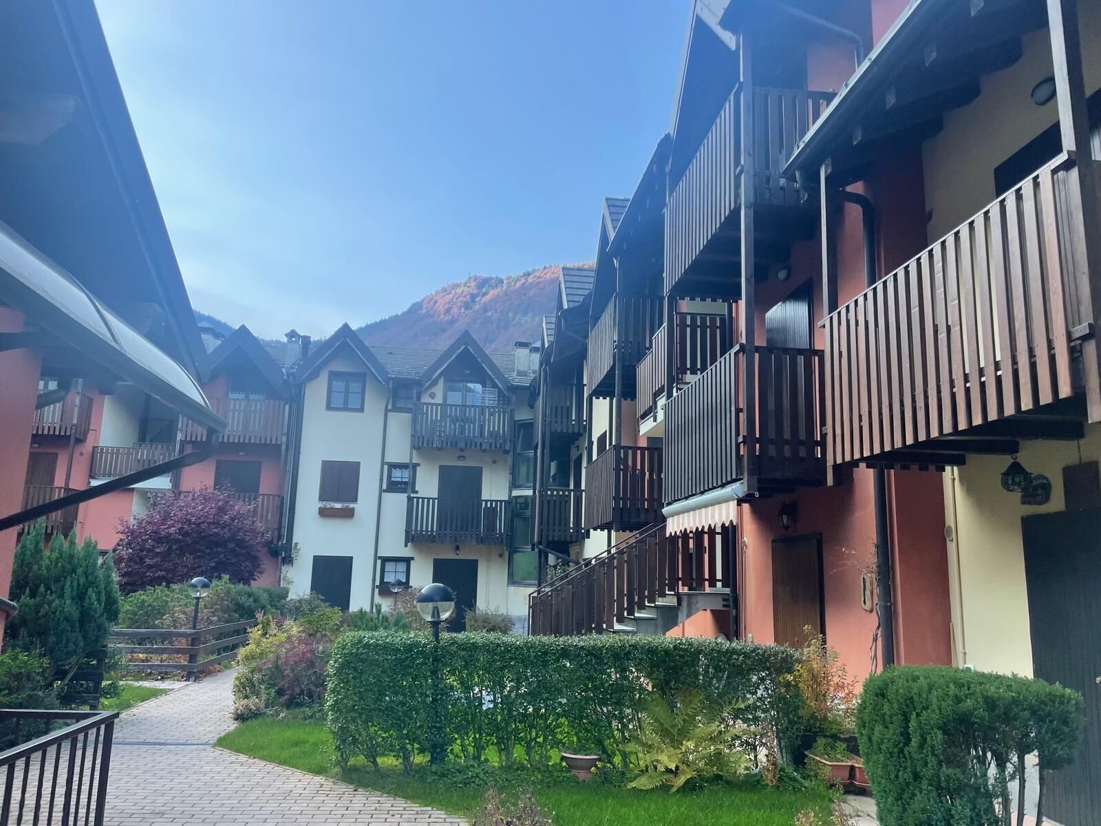 A narrow pathway leading to a row of multi-story buildings with balconies, surrounded by greenery and a mountain in the background.