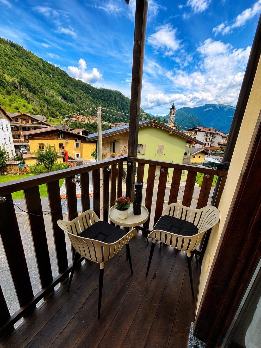 A balcony with two chairs and a small table, overlooking a mountainous village with traditional buildings.