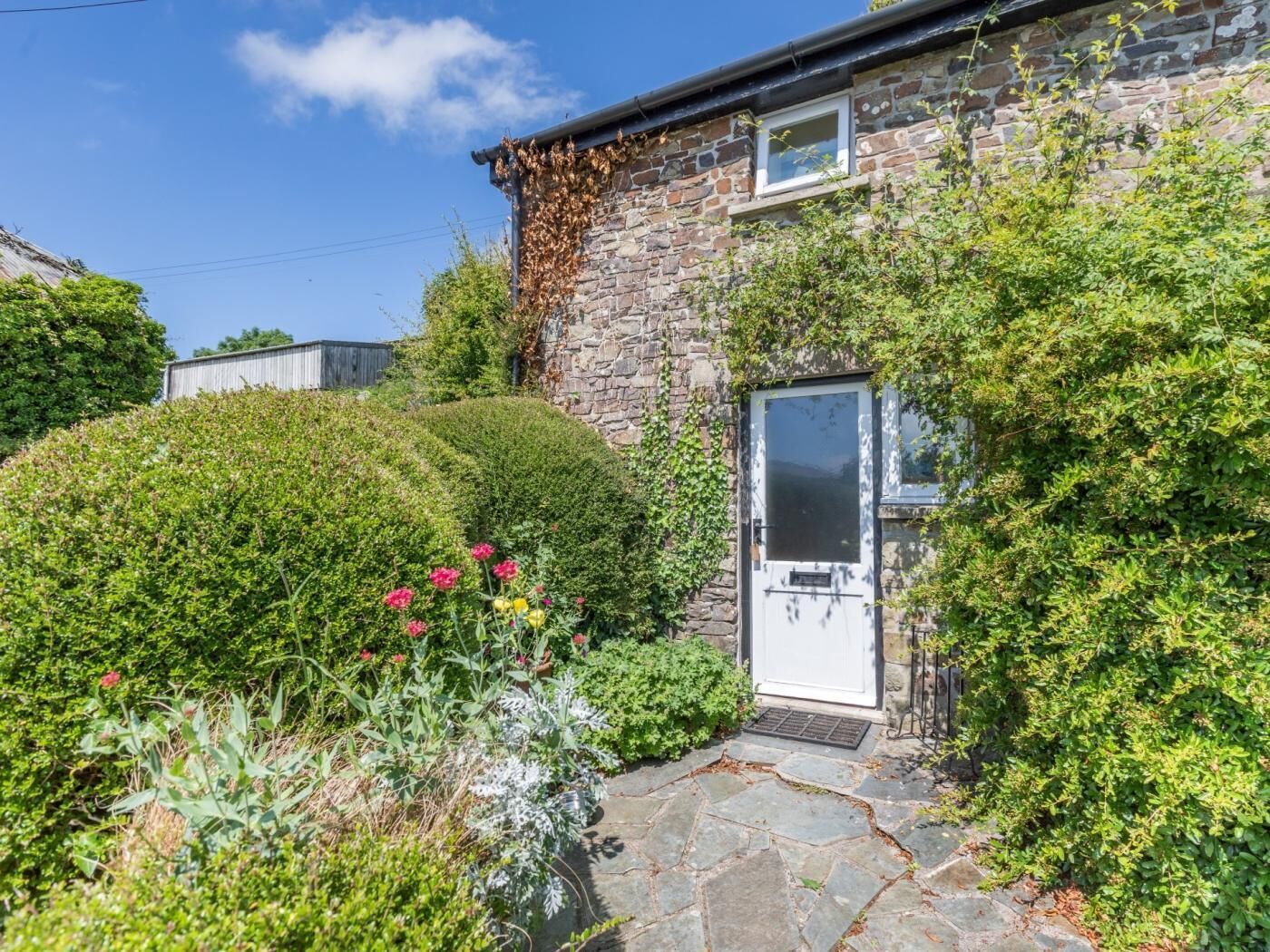 Une maison en pierre avec une porte blanche, entourée d’une végétation luxuriante et de plantes en fleurs.