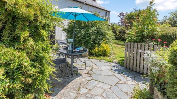 A patio with a table and chairs, a blue umbrella, and a stone pathway leading to a house. - 1 Bed in Okehampton (oc-42982) (Petrockstow)