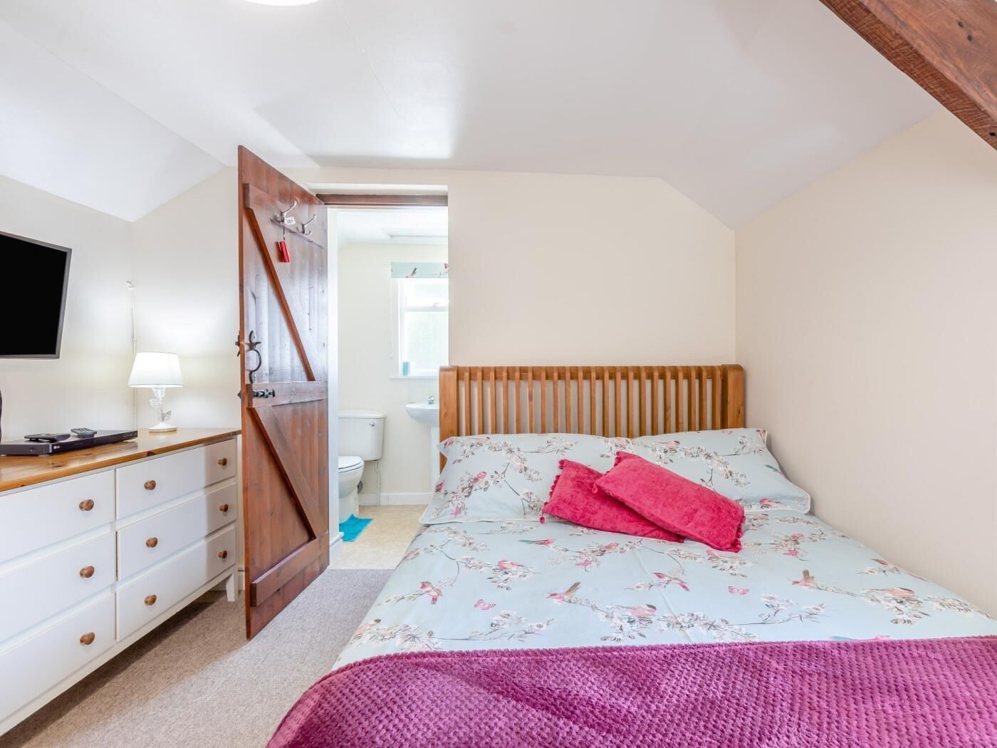 A bedroom with a bed, a dresser, a television, and a door leading to a bathroom.