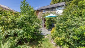 A stone pathway leading to a house with a blue umbrella, surrounded by dense greenery. - 1 Bed in Okehampton (oc-42982) (Petrockstow)