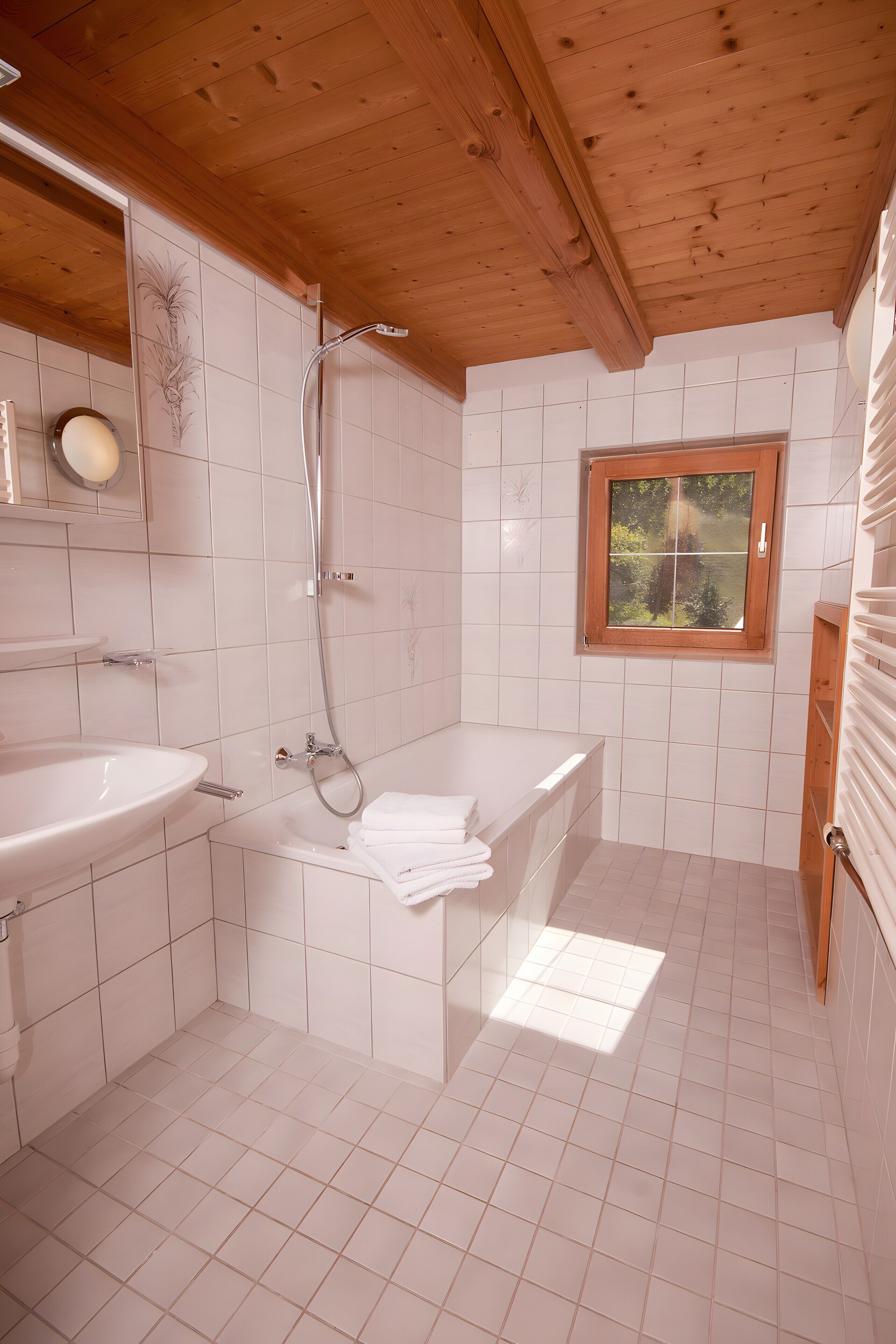 A bathroom with a wooden ceiling, white tiles, a bathtub, a sink, and a window with a view.