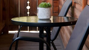 A wooden deck with a table and two chairs, a potted plant, and a view of greenery.