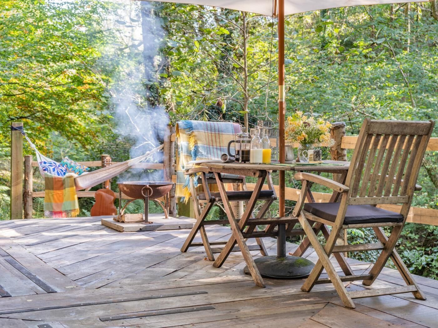 An outdoor patio with a wooden table, chairs, a hammock, and a grill.