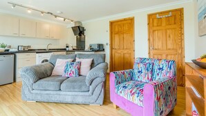 A compact living space with a kitchenette, a sofa, a colorful armchair, and wooden doors. - 1 Bed in Hayle (oc-43256) (Phillack)