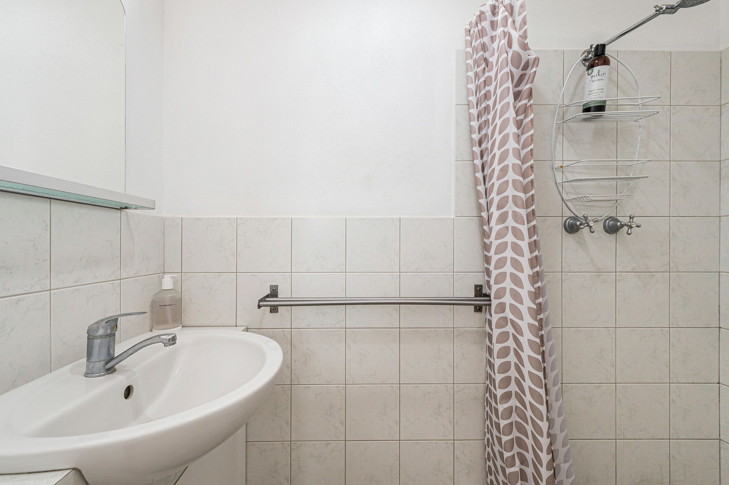 Une salle de bain avec un lavabo blanc, un porte-serviettes, un rideau de douche et une étagère en verre où sont rangés les articles de toilette.