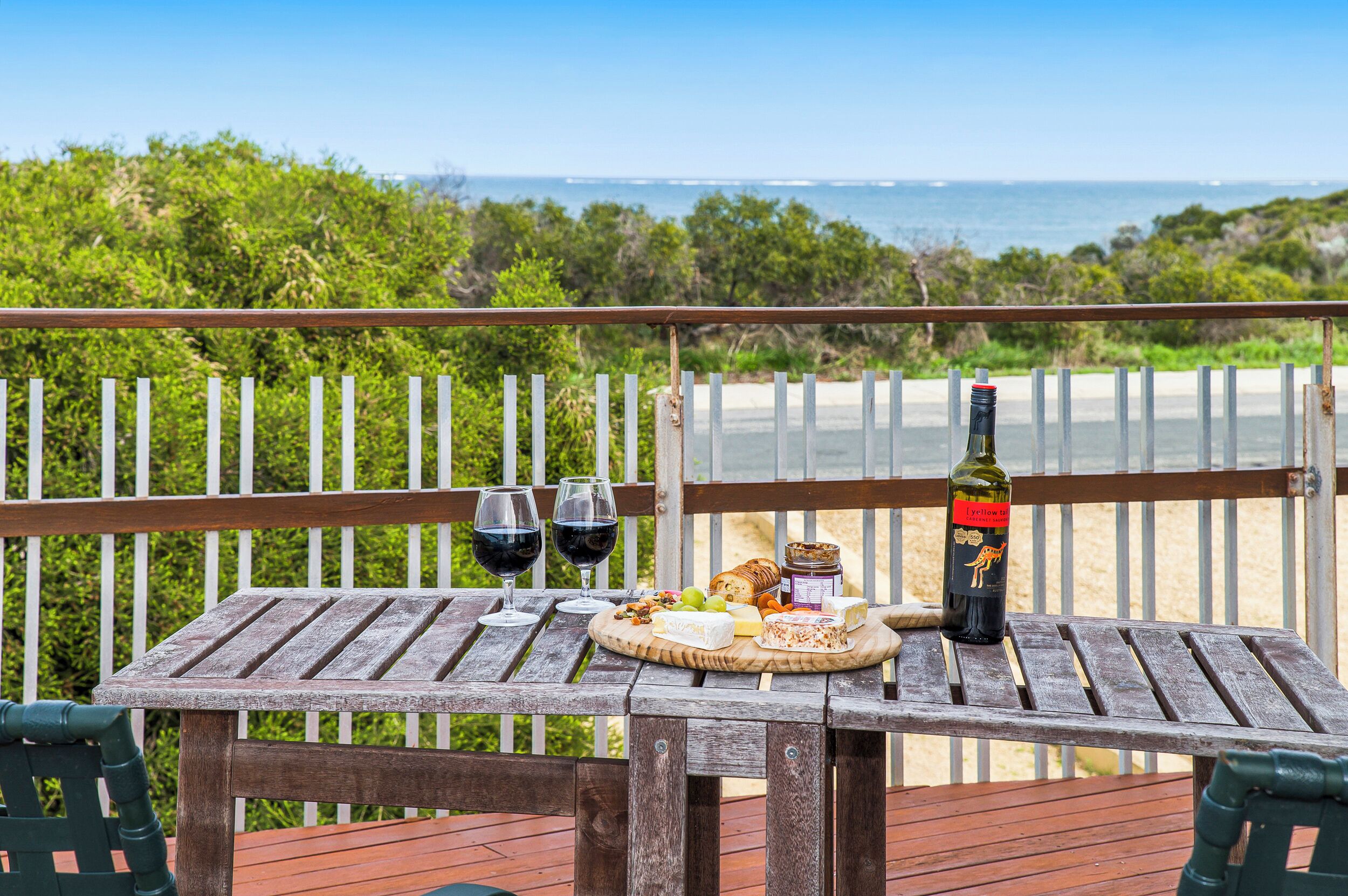 A wooden table with a bottle of wine, two glasses of red wine, a cheese platter, and a jar of jam on a balcony with a view of the ocean.