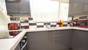 Una cocina moderna con encimera blanca, armarios grises, un lavamanos de acero inoxidable y una tetera roja.