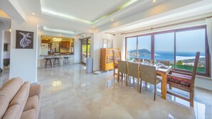 Un vaste espace de vie comprenant une table à manger, des chaises, un canapé, une cuisine en arrière-plan et une grande fenêtre donnant sur la mer.