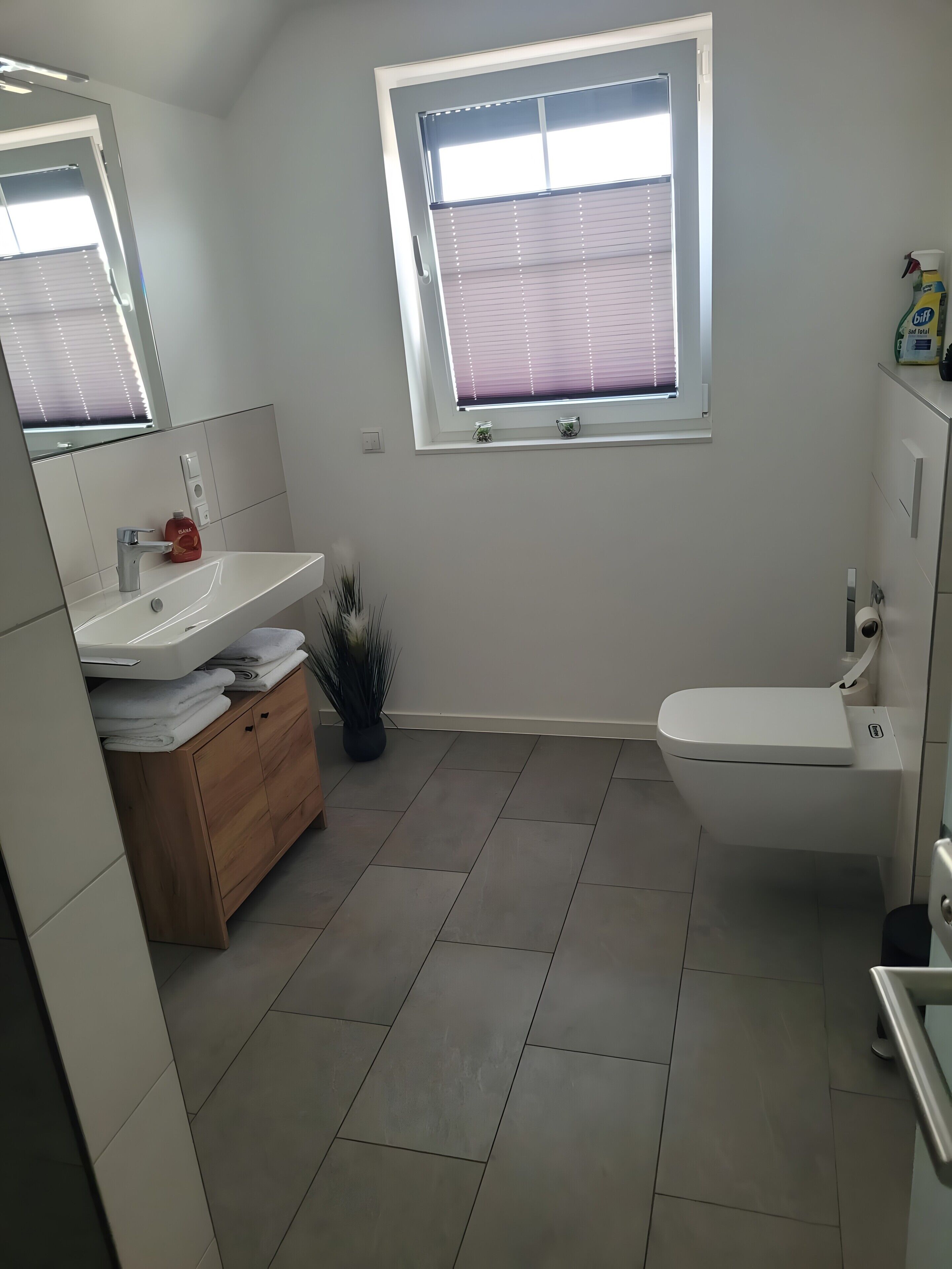 A modern bathroom with a white sink, mirror, and toilet. There is a window with blinds, a small plant, and a wooden cabinet.