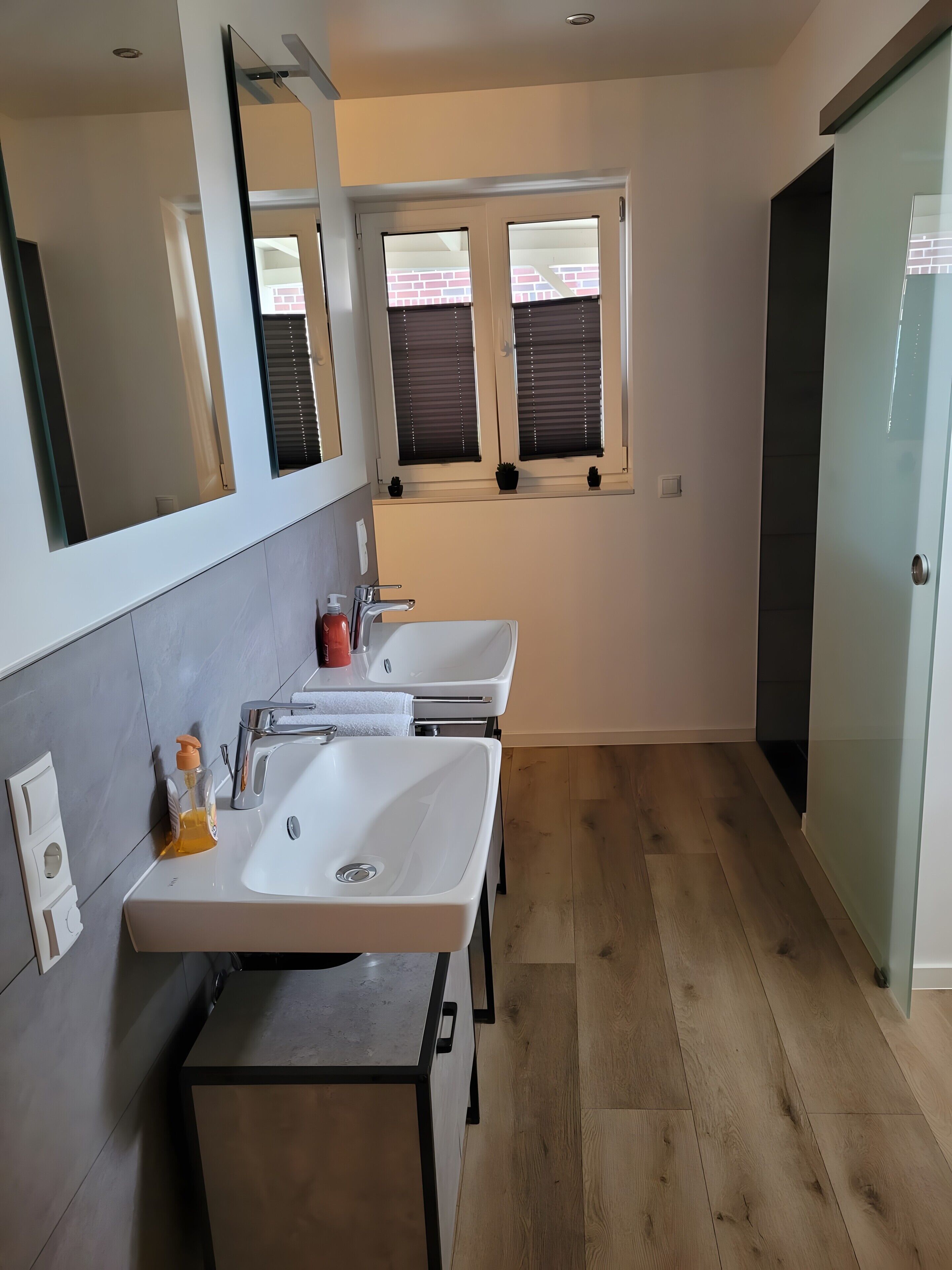 Ein modernes Badezimmer mit zwei Waschbecken, einem Spiegel und einem Fenster mit Jalousien.