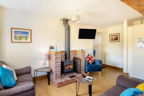 A living room with a brick fireplace, a blue armchair, a small table with a glass of wine, and a TV mounted on the wall. - 3 Bed in Whitchurch Canonicorum (oc-43398) (Whitchurch Canonicorum)