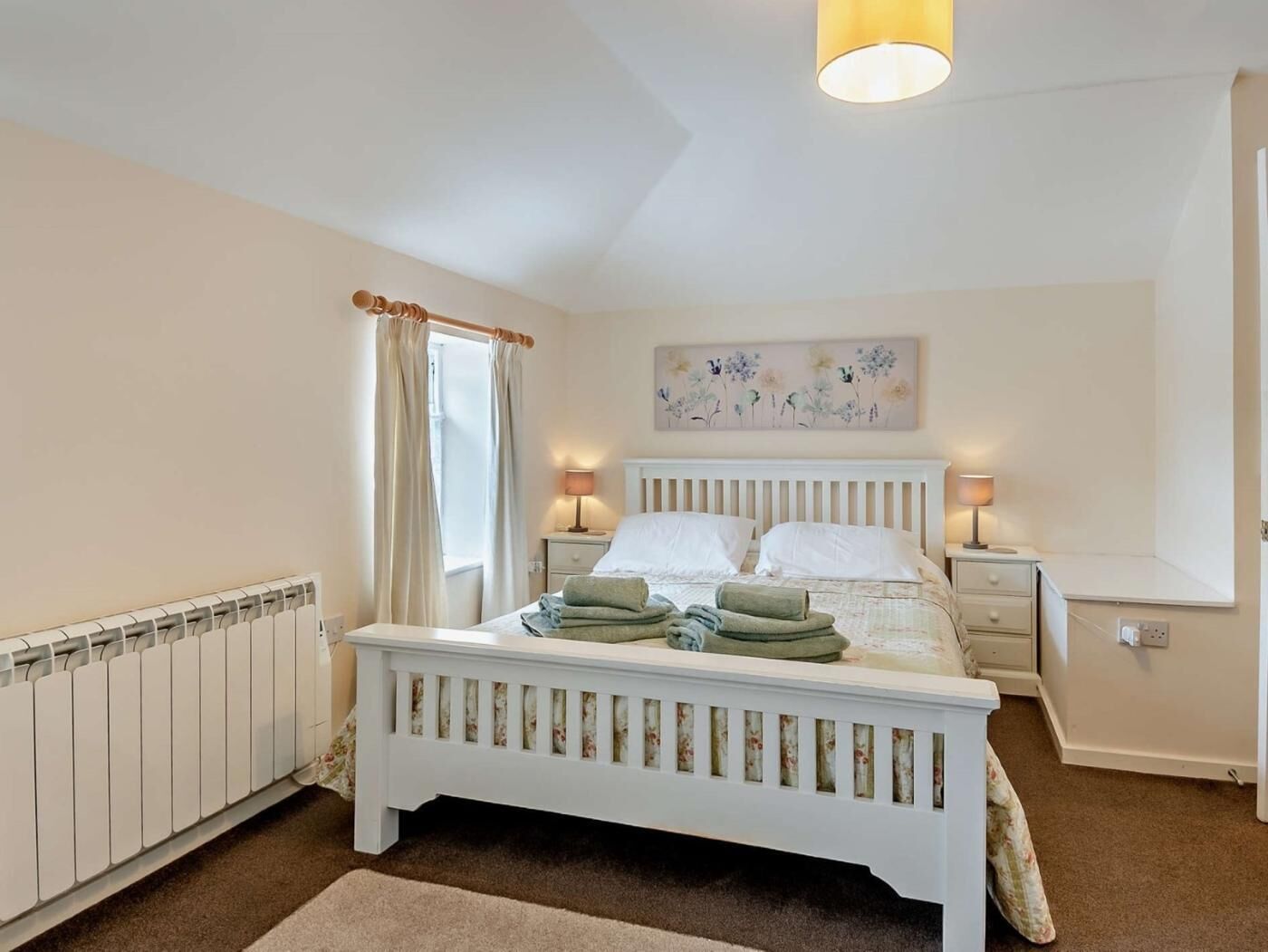 A bedroom with a white bed, two bedside lamps, a radiator, and a window with curtains.