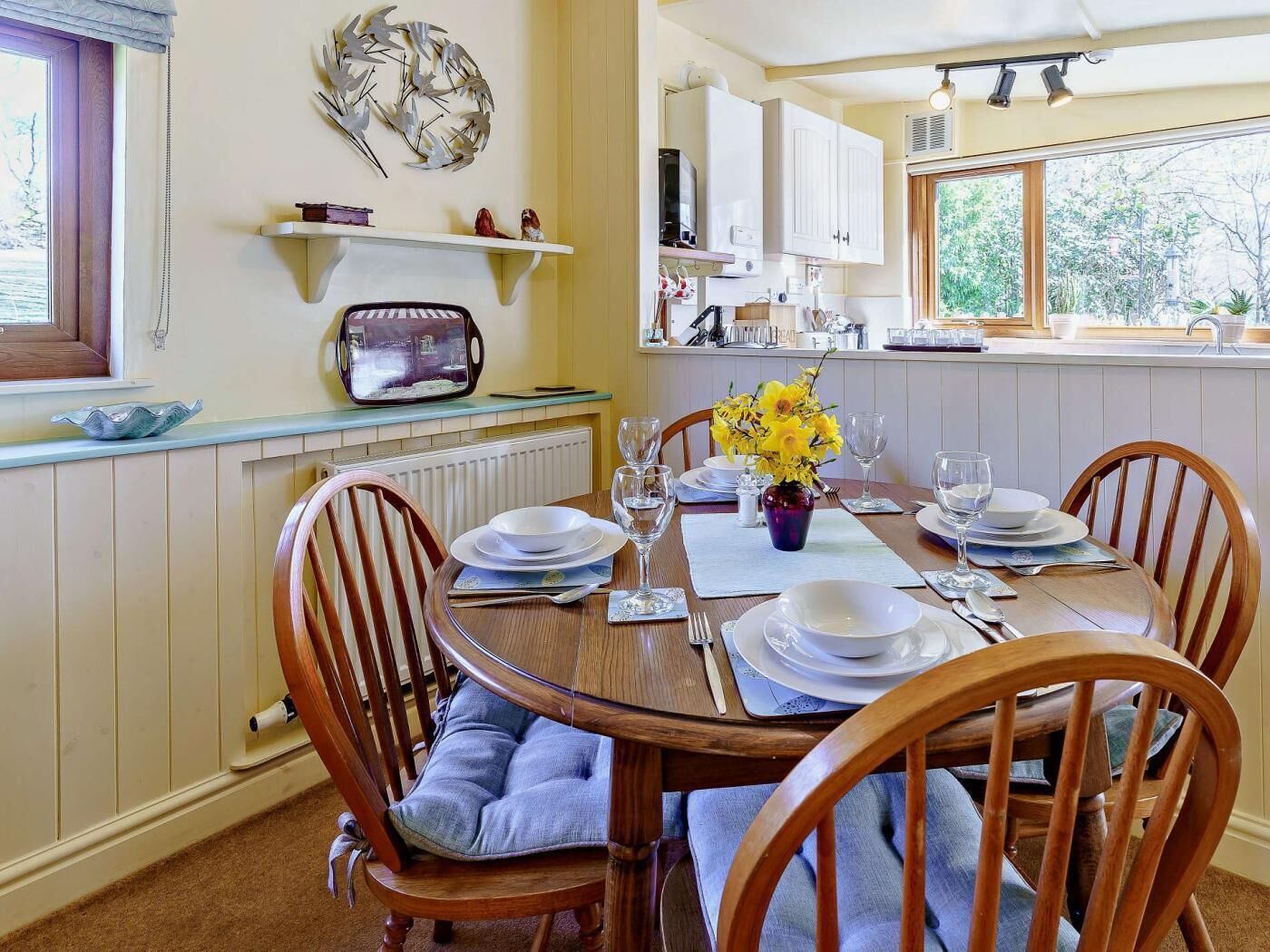 A dining area with a wooden table set for four, chairs with blue cushions, a vase with yellow flowers, and a kitchen in the background.