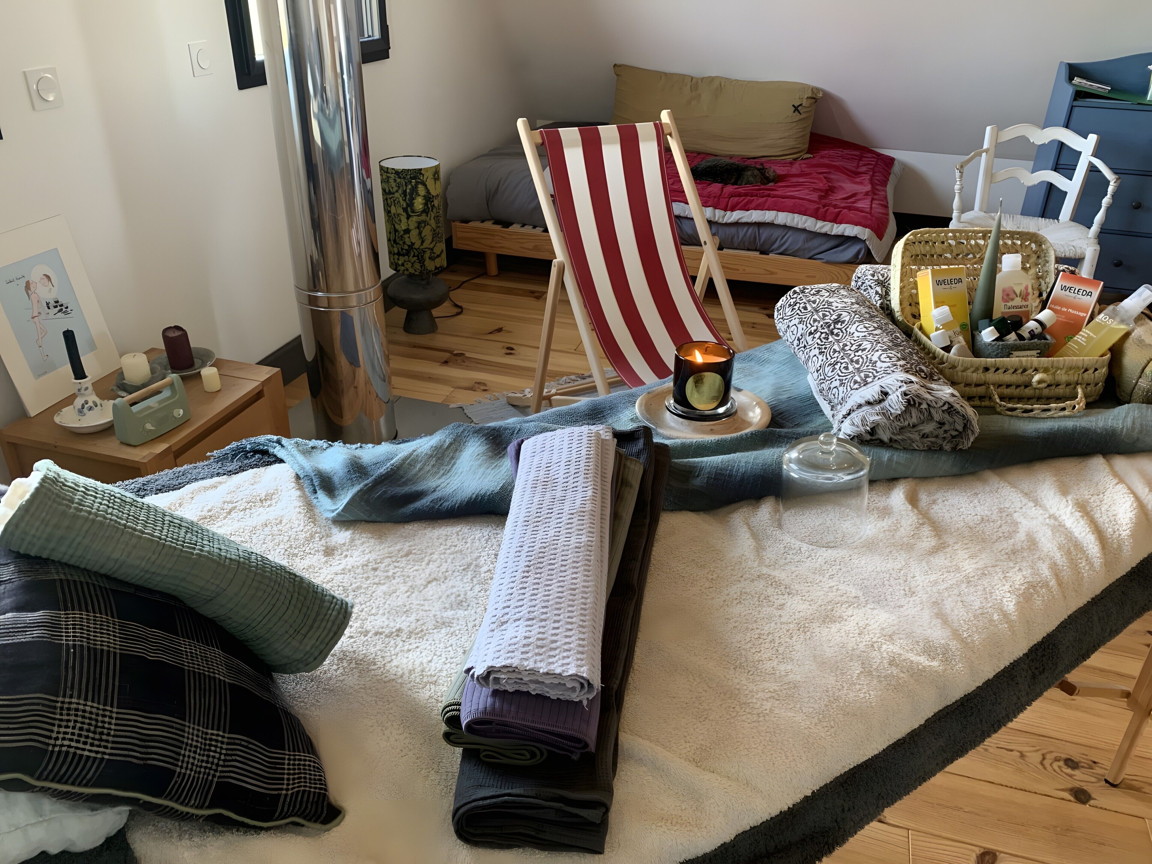 Un lit bien fait, agrémenté d’une chaise de plage à rayures, d’une bougie allumée et d’un panier rempli de produits de toilette.