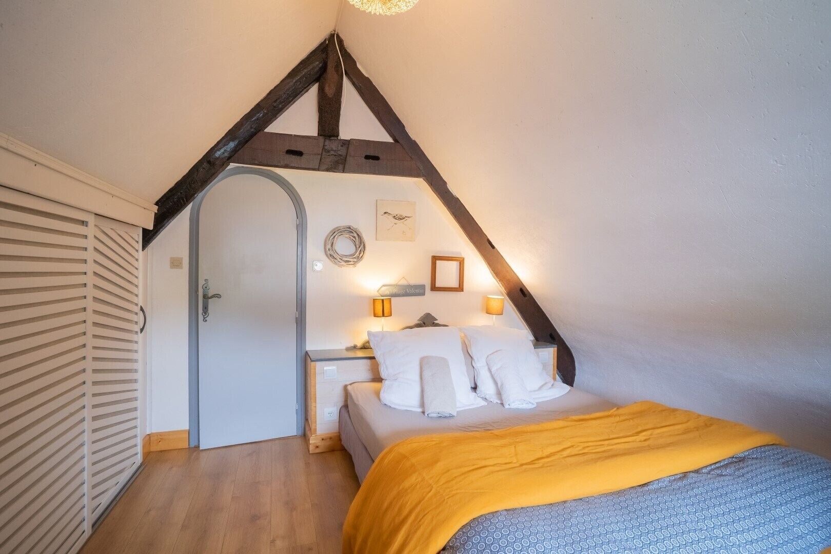 Un dormitorio con techo inclinado, una cama con ropa de cama blanca, una manta amarilla y una mesita de noche pequeña con una lámpara.