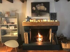 A fireplace with a lit fire, a bookshelf with various items, a framed painting above the fireplace, and a sign with text.