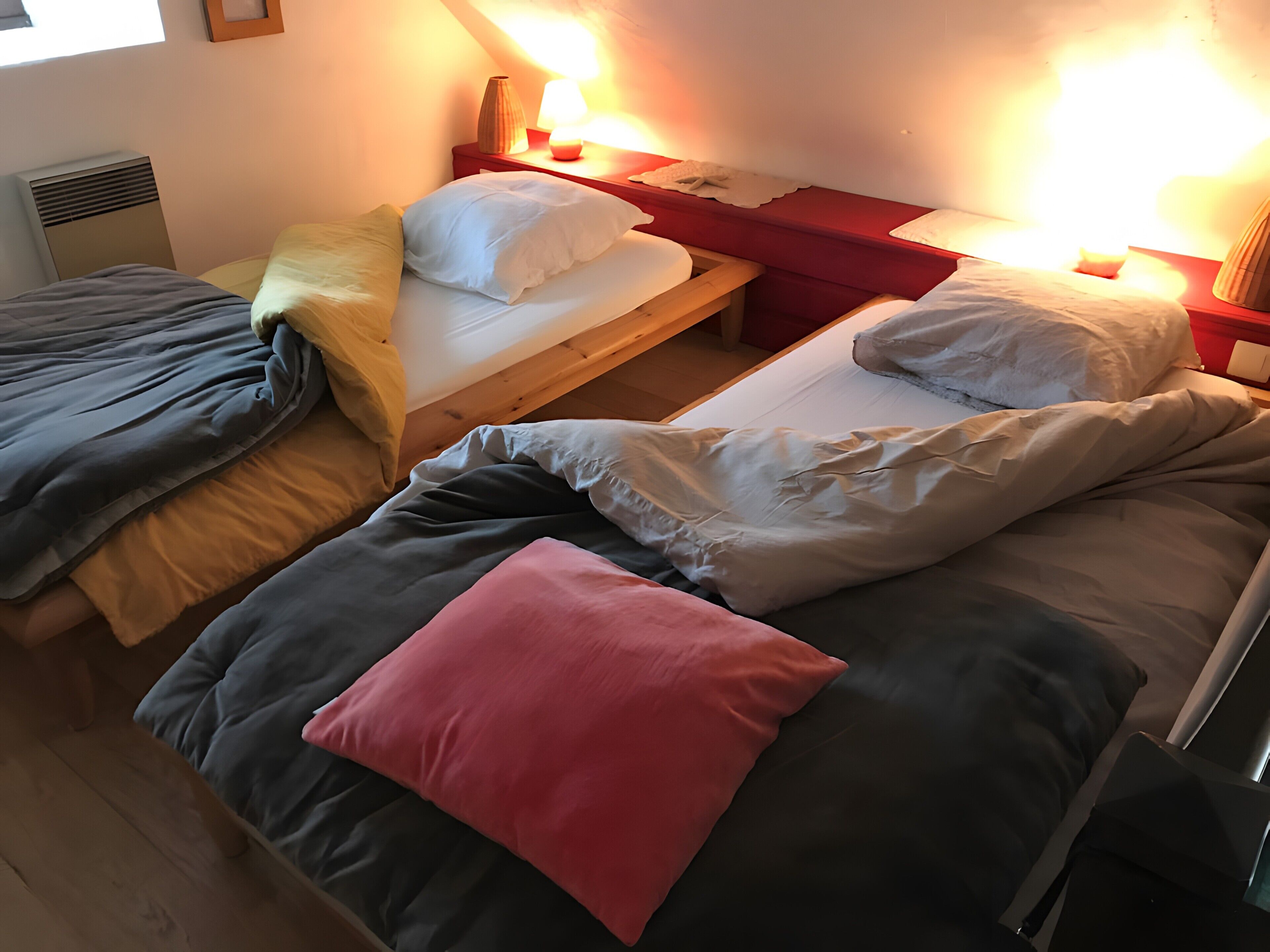 A room with three beds, each with different bedding colors and a bedside table with a lamp.