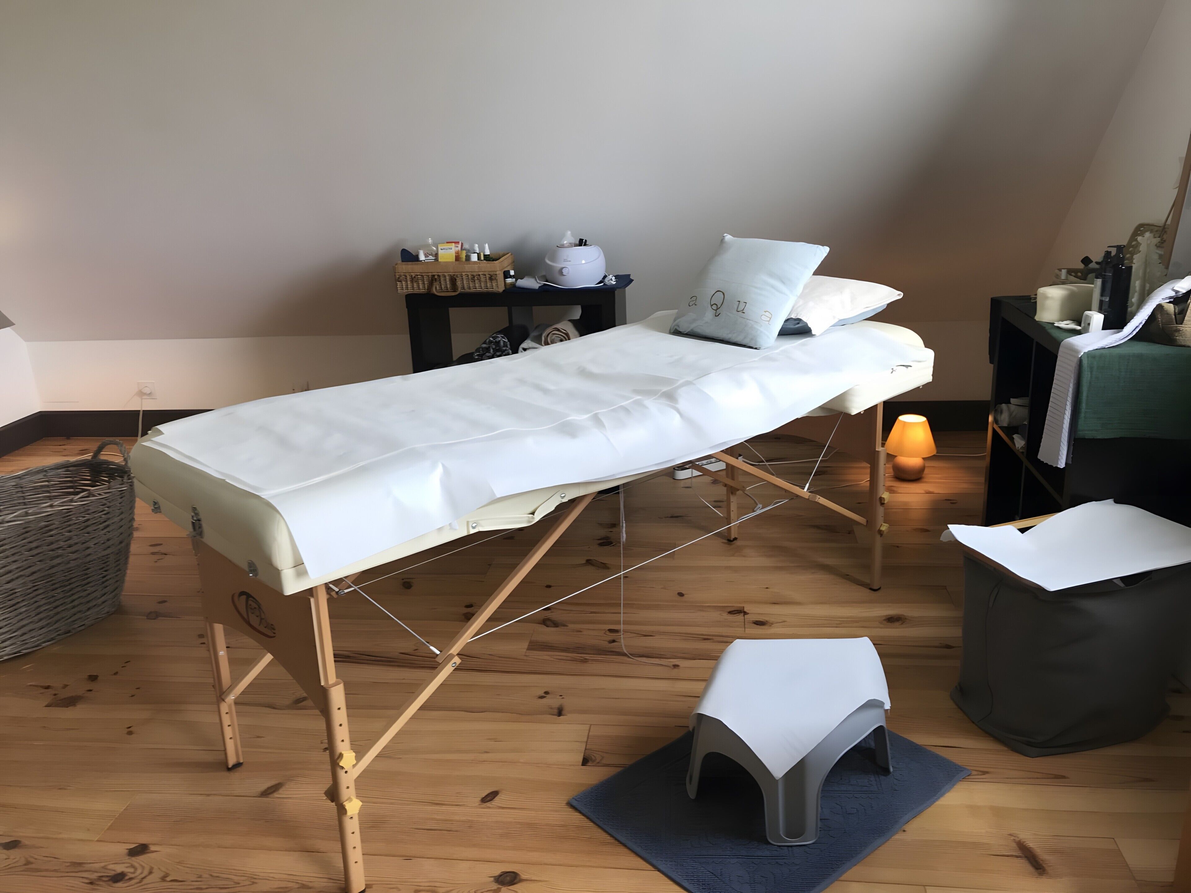 Une pièce avec un plancher en bois, une table de massage recouverte d’un drap blanc, un petit tabouret, un panier et une table de chevet avec une lampe.