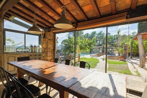 A wooden dining table with chairs, a stone wall, and a view of a swimming pool and garden.