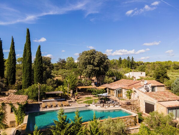A pool area with a clear blue pool, surrounded by stone buildings, cypress trees, and a hillside backdrop.