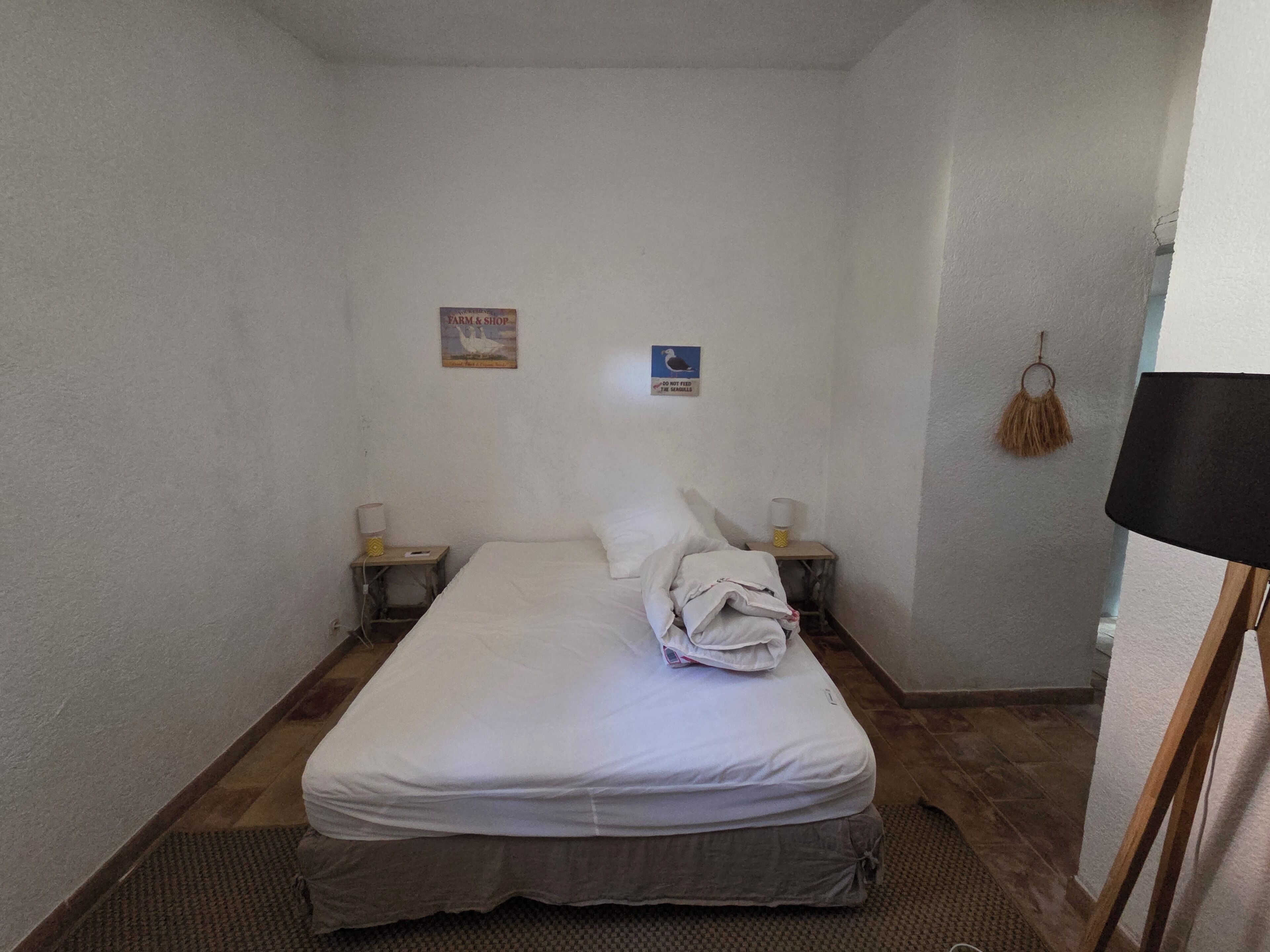 A small, simple room with a single bed, two bedside tables, a lamp, and a wall-mounted hanging.