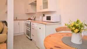 A compact kitchen with white cabinets, a microwave, and a small oven. There's a dining table with a vase of flowers and a chair. - 1 Bed in Bude (oc-43697) (Morwenstow)