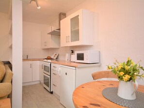 Eine kompakte Küche mit weißen Schränken, einer Mikrowelle und einem kleinen Backofen. Ein Esstisch mit Blumenvase und einem Stuhl stehen bereit.