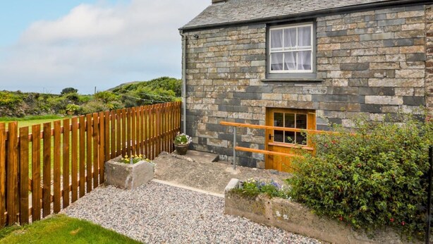 A stone house with a wooden door, a window, and a gravel pathway leading to a wooden fence. - 1 Bed in Bude (oc-43697) (Morwenstow)