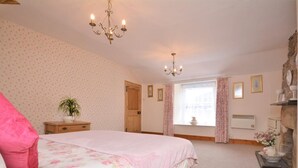 A bedroom with a bed, a chandelier, a window with curtains, a radiator, and a small table with a plant. - 1 Bed in Bude (oc-43697) (Morwenstow)
