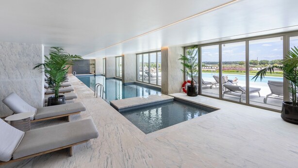 Un área de piscina interior moderna con vistas al exterior, suelo de mármol y sillones de descanso.